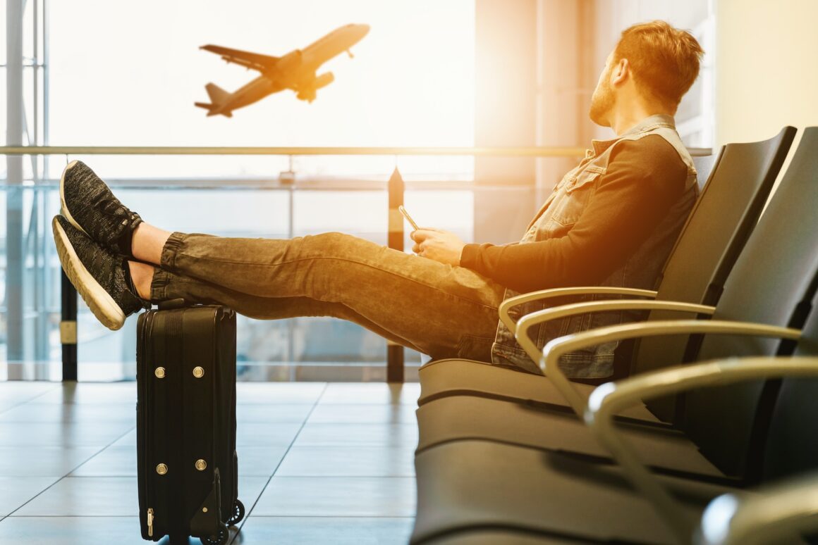 man sitting on gang chair with feet on luggage looking at airplane long layover