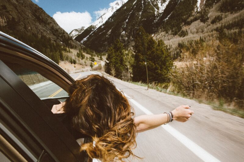 woman riding on vehicle putting her head and right arm outside the window while travelling the road Road Trip Ideas