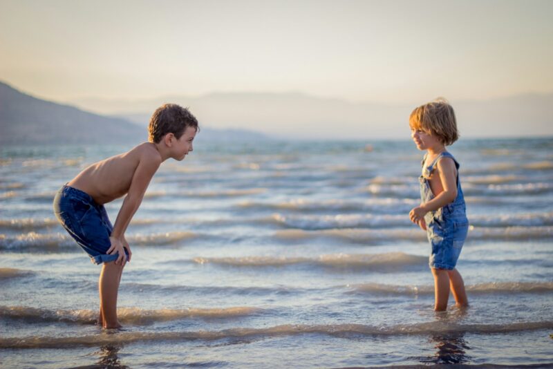 boy bending knee while watching younger child on beach, family travel tips